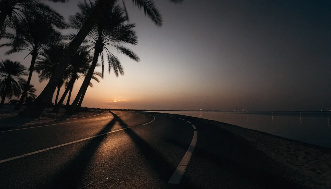 A coastal Gulf carriageway at dusk with long shadows crossing the lanes and warm light on the horizon